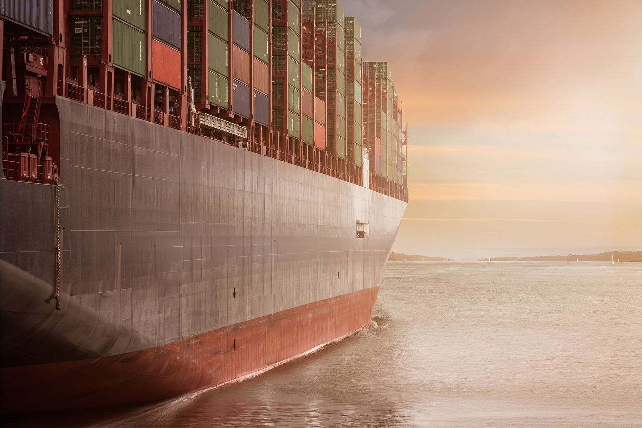 About A colossal cargo ship loaded with containers navigates through calm waters against a vibrant sunset sky.