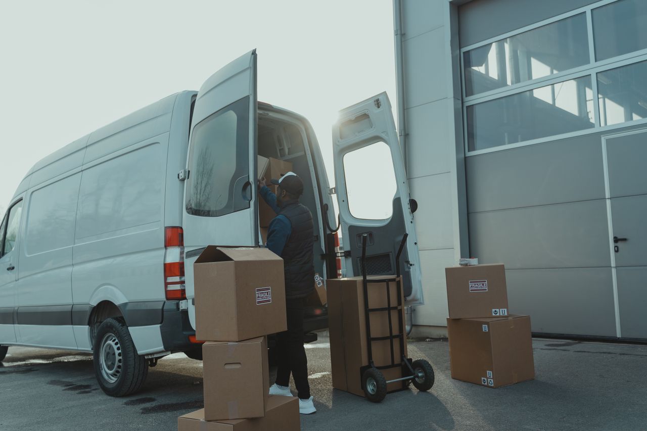 Services Courier loading cardboard boxes into a delivery van outside a warehouse.